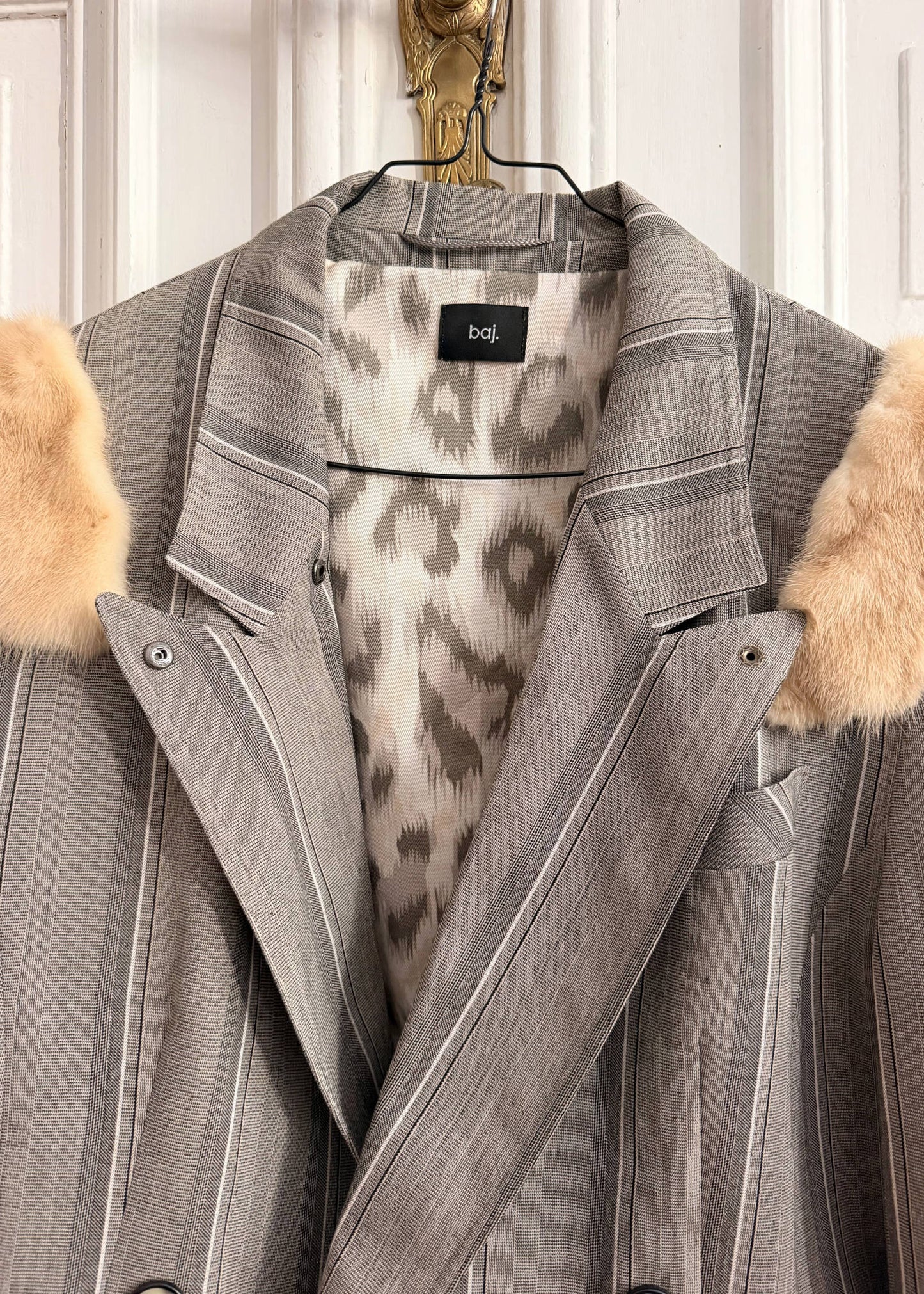 A gray pinstripe blazer jacket with beige faux fur trim on the collar and shoulders, featuring a leopard-print lining and a black 'bdt.' label at the inner collar, hanging on a black hanger against a white paneled door.