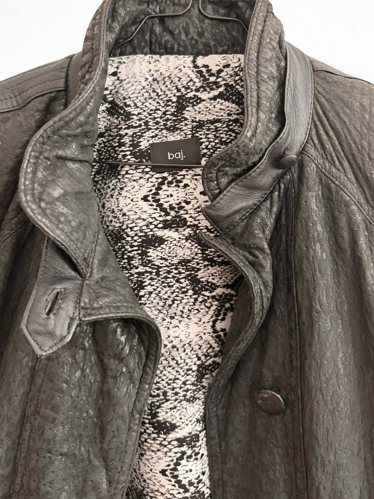 A close-up of a dark gray textured leather jacket with a high collar, worn over a black and beige snakeskin-patterned shirt. The brand label 'b&j' is visible inside the collar.