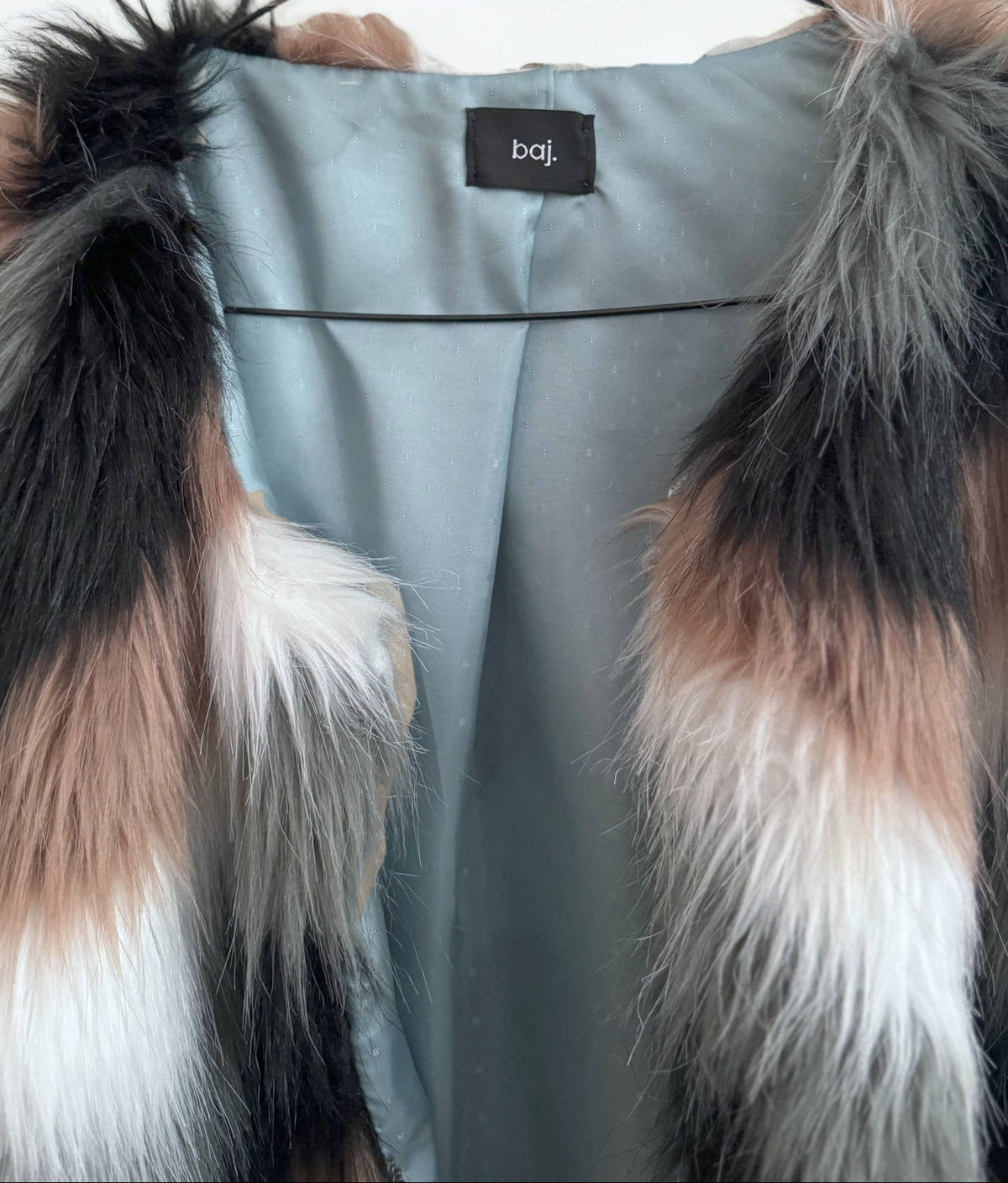 A close-up of a faux fur vest with a color-blocked pattern of black, brown, white, and gray, worn over a dusty blue satin inner layer. A black brand label reading 'baj.