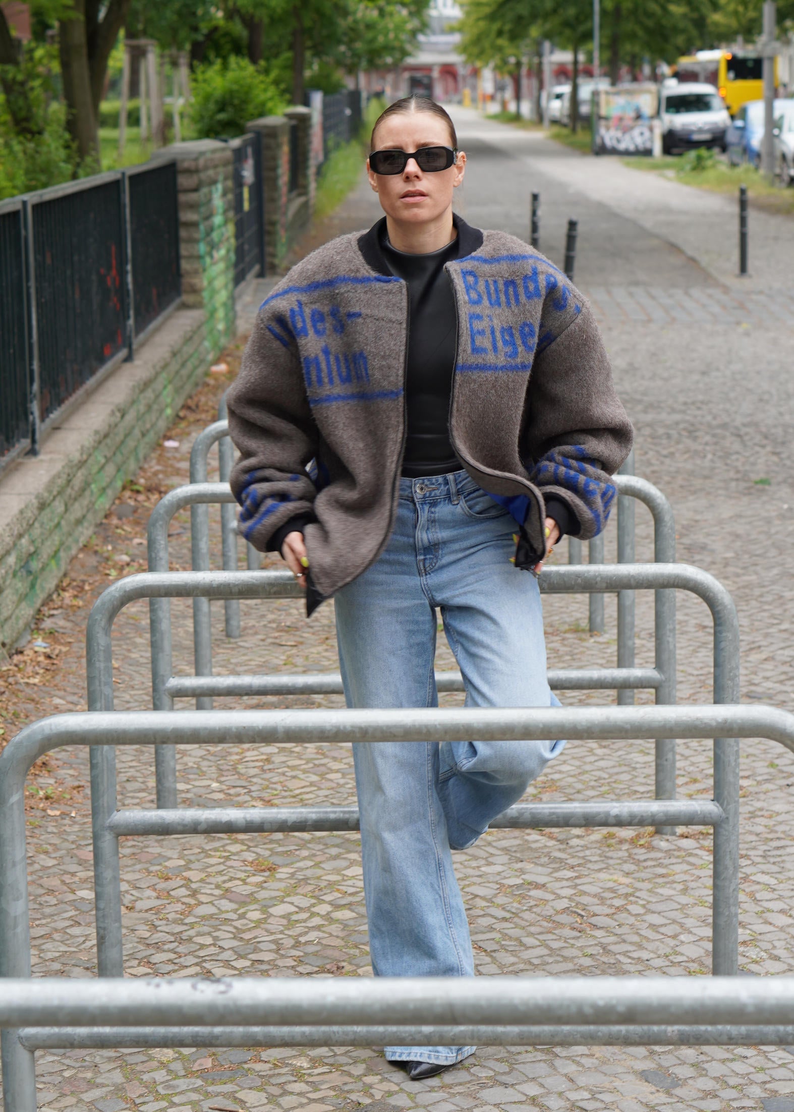 A woman wearing a brown knit cardigan with blue lettering and light blue jeans, standing on a cobblestone street behind metal bike racks.