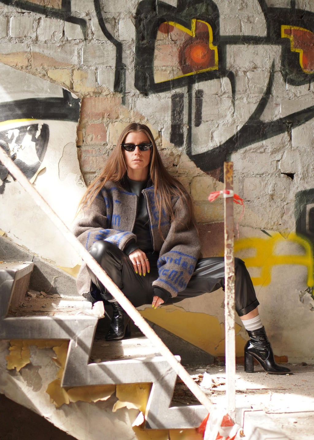 A woman wearing a brown textured jacket with blue and yellow graphic details, black pants, and black boots sits on concrete stairs in front of a graffiti-covered brick wall.