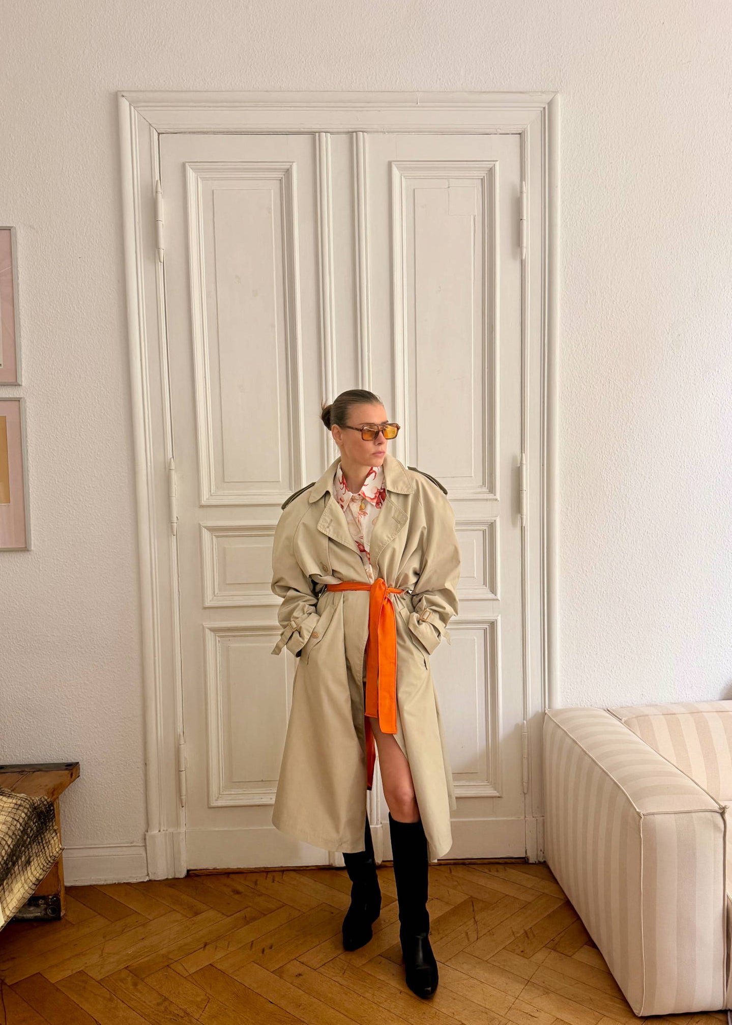 Person wearing a beige trench coat and orange skirt in a room with white walls and wooden floor.