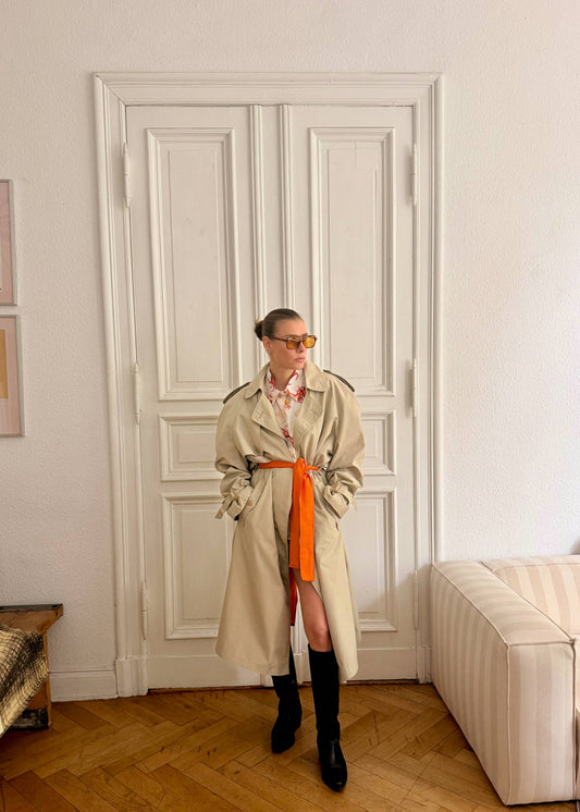 Person wearing a beige trench coat and orange skirt in a room with white walls and wooden floor.