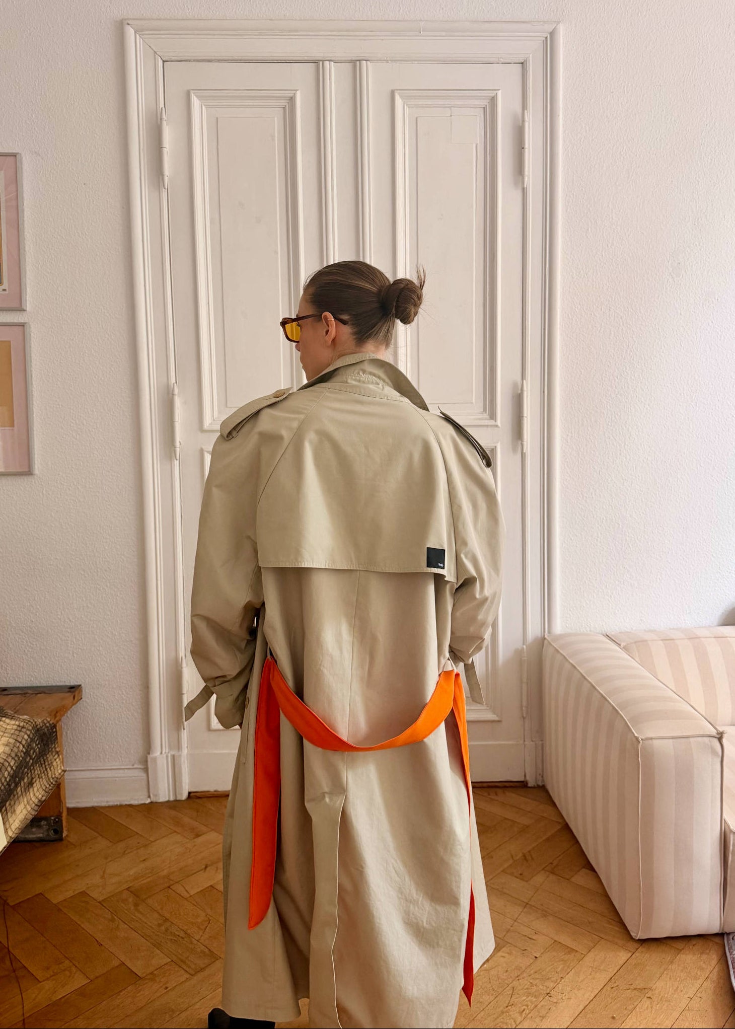 Person wearing a beige trench coat with an orange belt in a room with a white door and light-colored furniture.