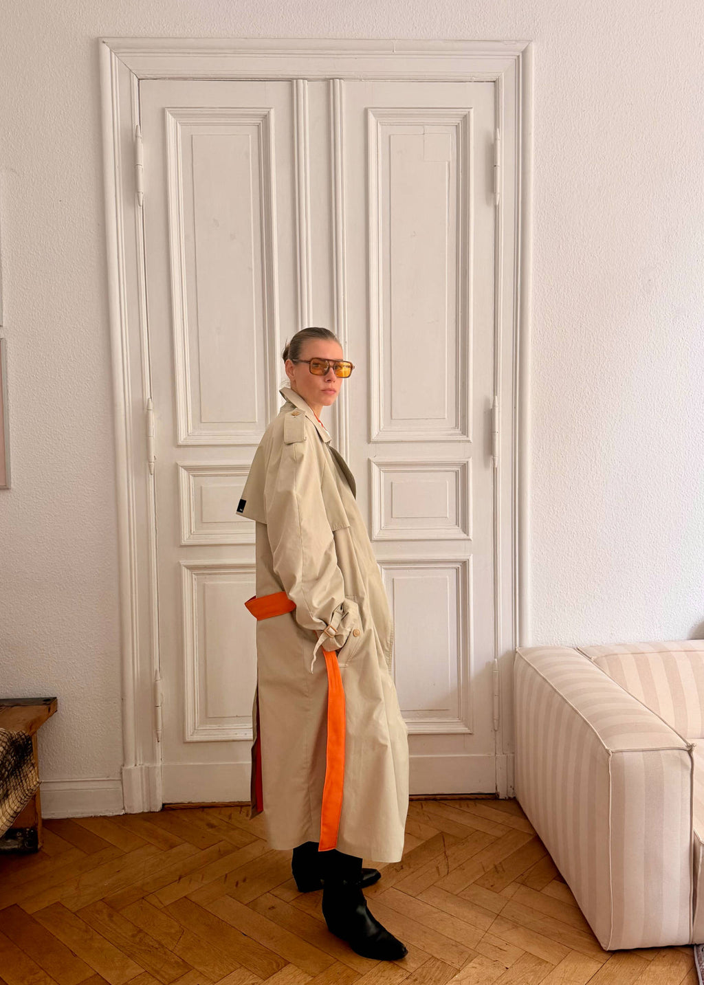 Person wearing a beige trench coat with an orange sash in a room with white walls and wooden flooring.