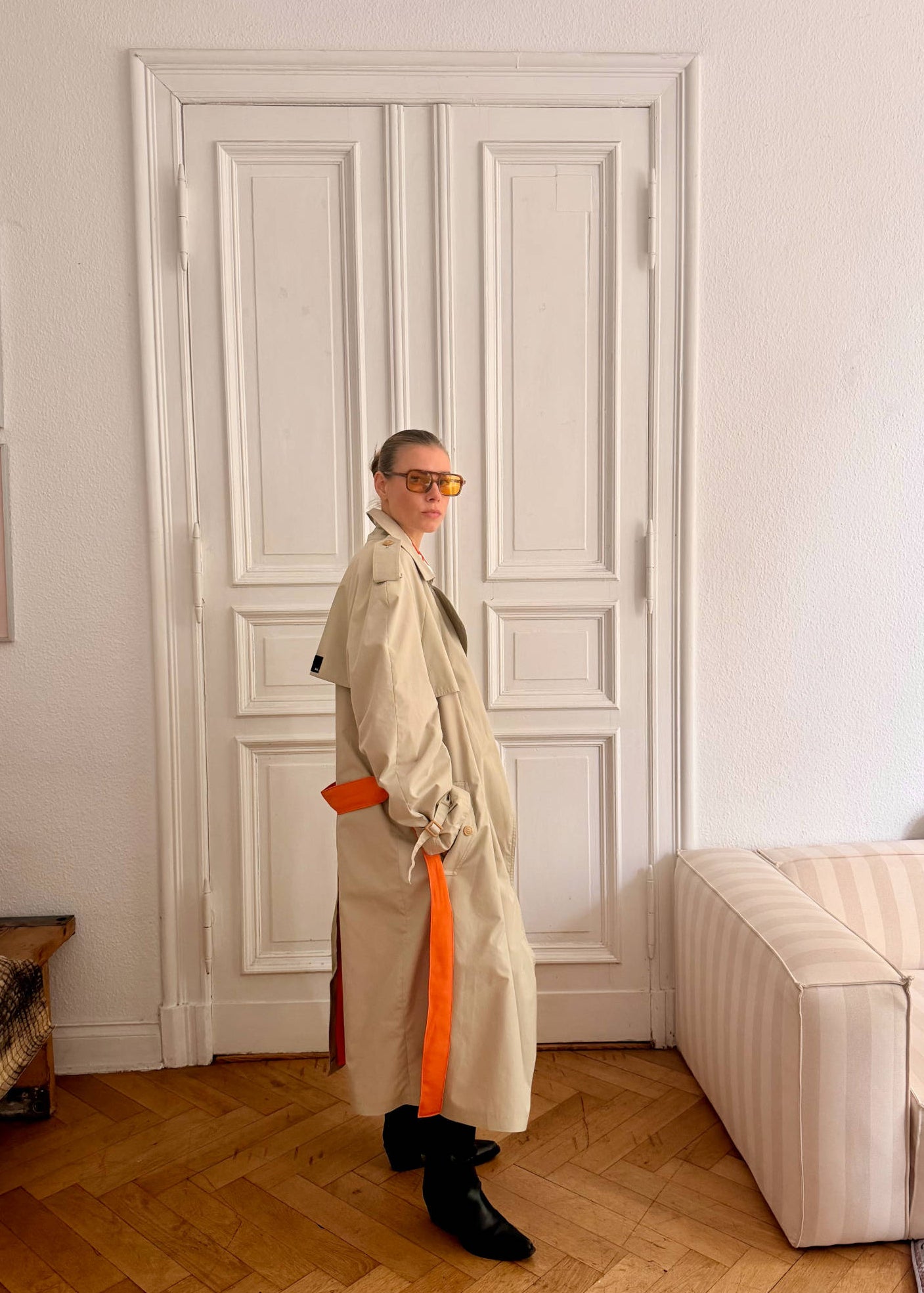 Person wearing a beige trench coat with an orange sash in a room with white walls and wooden flooring.