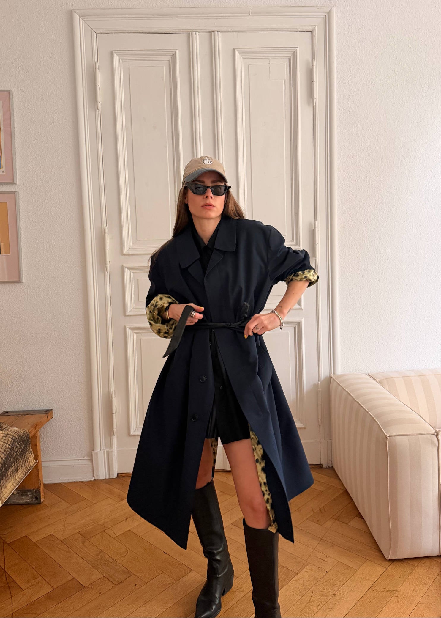 Woman in a stylish outfit with a long vintage trench coat and knee-high boots in a living room.