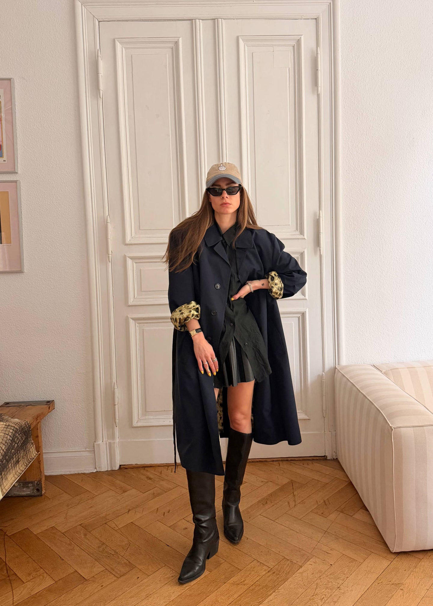Woman in a long dark coat standing in a room with a white door and beige sofa.