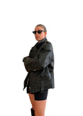 A woman wearing a black leather jacket, black shorts, and black sunglasses stands with arms crossed in a minimalist room with a wooden floor and a cream-colored sofa.