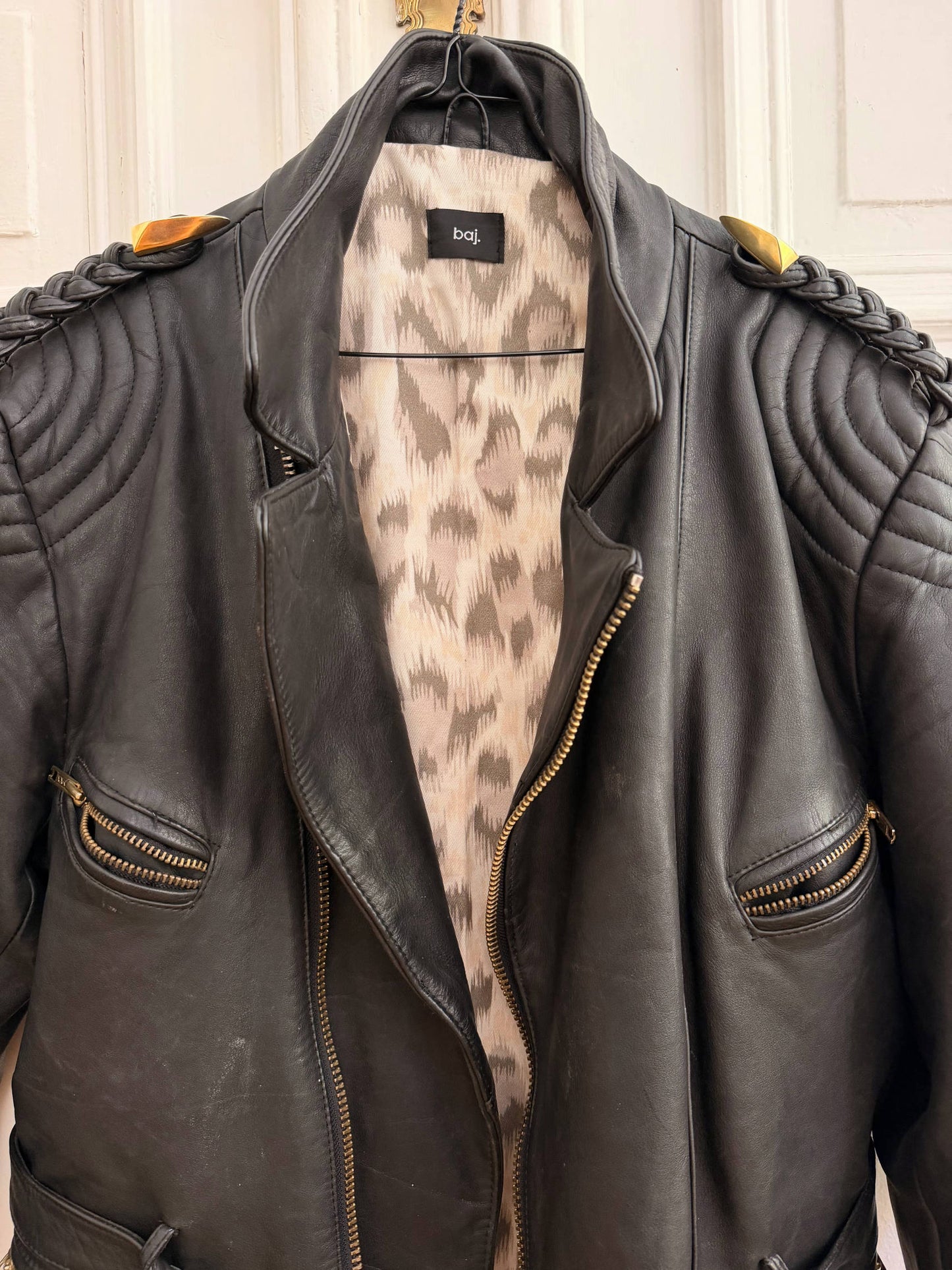 A black leather motorcycle jacket with quilted shoulder details and a leopard-print lining, hanging on a hanger against a white paneled door.