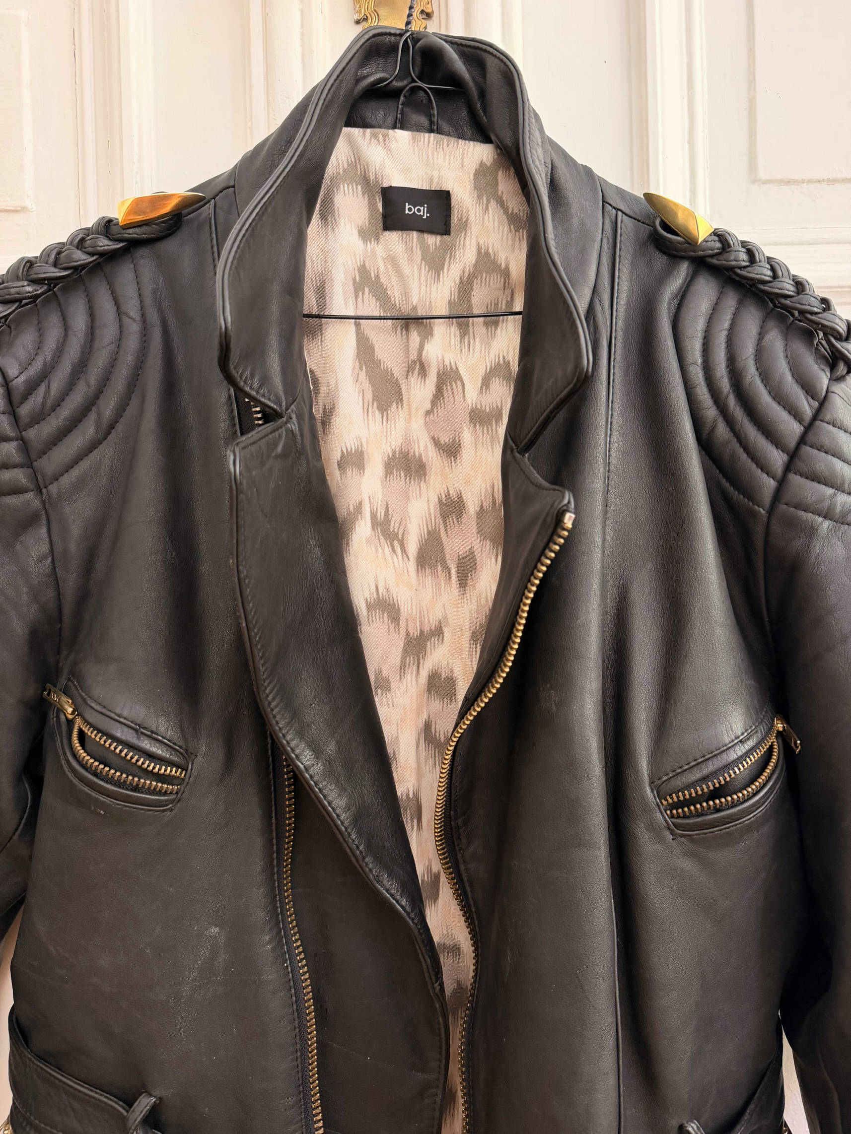 A black leather motorcycle jacket with quilted shoulder details and a leopard-print lining, hanging on a hanger against a white paneled door.