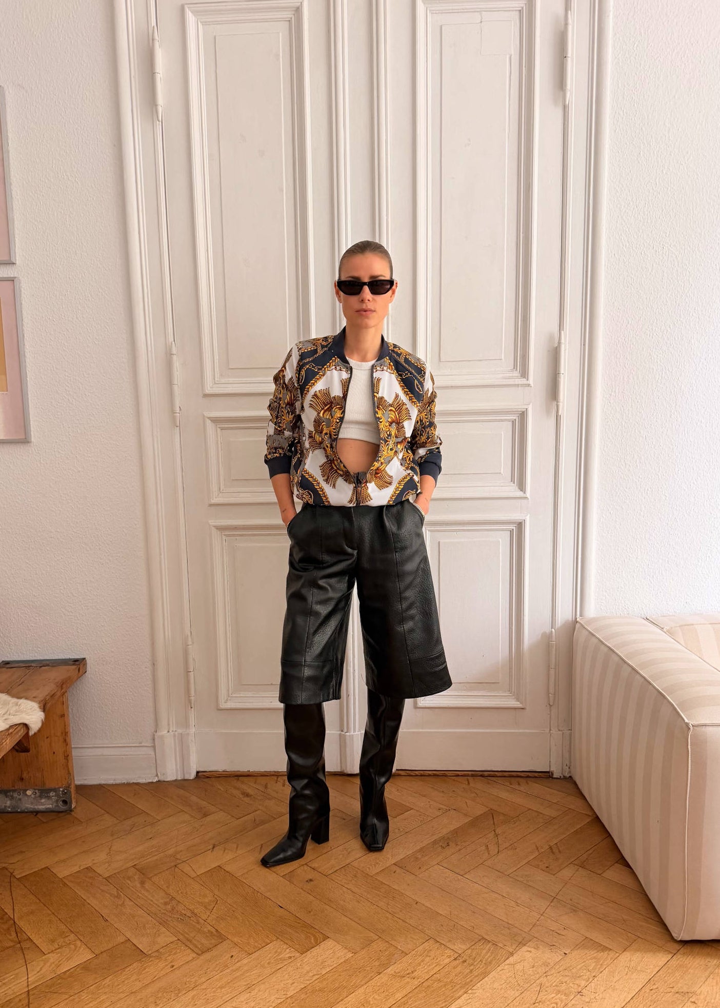 A person stands indoors in front of white paneled double doors on a wood parquet floor, wearing a patterned cropped jacket over a white cropped top, black leather shorts, and black knee-high boots.