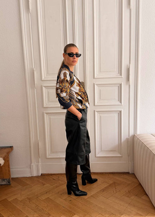 A person wearing a patterned shirt, black trousers, black boots, and sunglasses stands in a bright room with white paneled doors, a wooden floor, and a cream sofa.