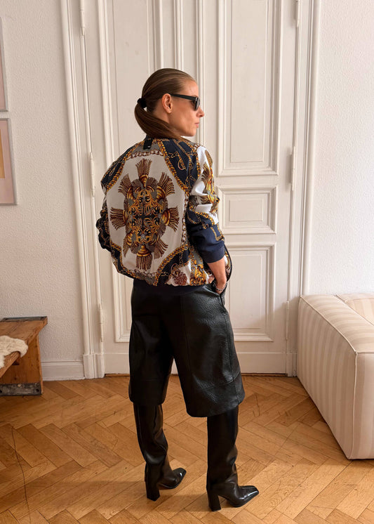 A person stands in a room with wood floors and white paneled doors, wearing a patterned cropped jacket with a large ornate back motif, black leather-like pants, and black boots.
