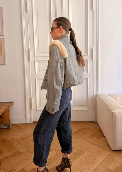 A woman stands in profile in a bright room with white walls, a paneled door, wood flooring, and a cream sofa. She is wearing a light gray cropped jacket with a beige shearling-style collar, dark blue jeans, and brown lace-up ankle boots.