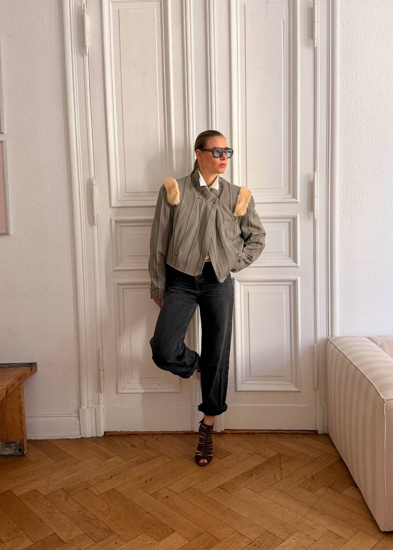 A person stands in a bright room with white paneled doors, wearing a gray cropped jacket with beige shoulder details, a white collared shirt, dark jeans, and brown shoes.