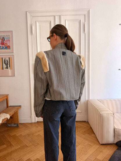 A woman viewed from behind wearing a gray striped cropped jacket with beige fur-like shoulder patches and dark blue jeans, standing in a bright room with a white door, wood floor, and cream sofa.