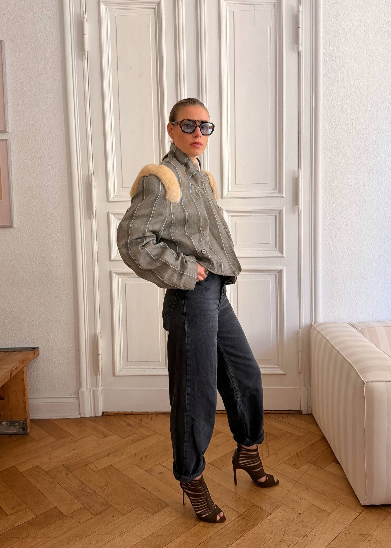 A person stands in a bright indoor room wearing a gray striped jacket with a beige fur-like collar, dark wide-leg jeans, and brown heeled ankle boots. The room has white paneled doors, a light wood floor, framed wall art, and a cream sofa.