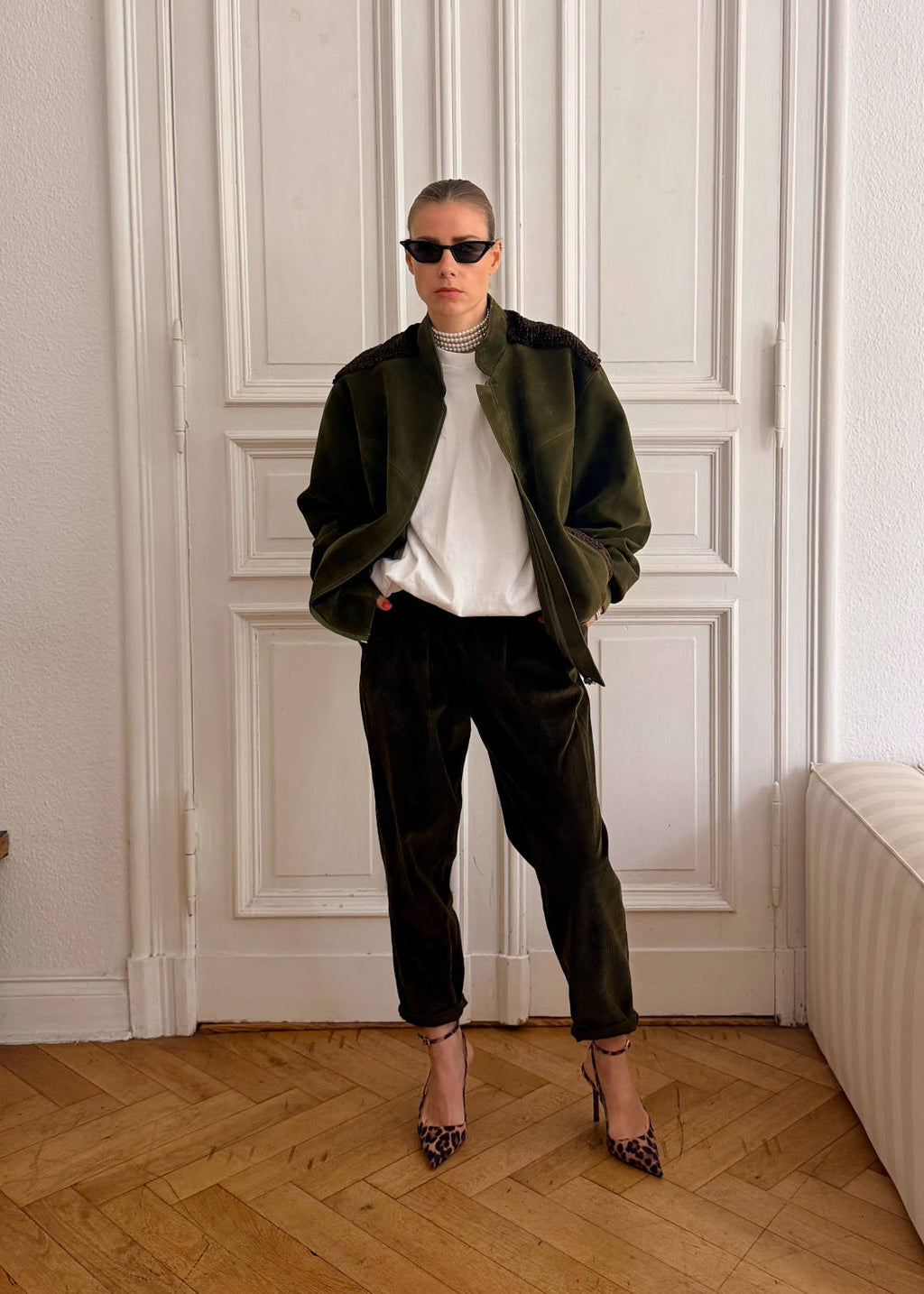 A person stands in a bright room with white paneled doors, a wood herringbone floor, and a cream sofa, wearing an olive green jacket over a white top, dark trousers, and leopard-print heels.