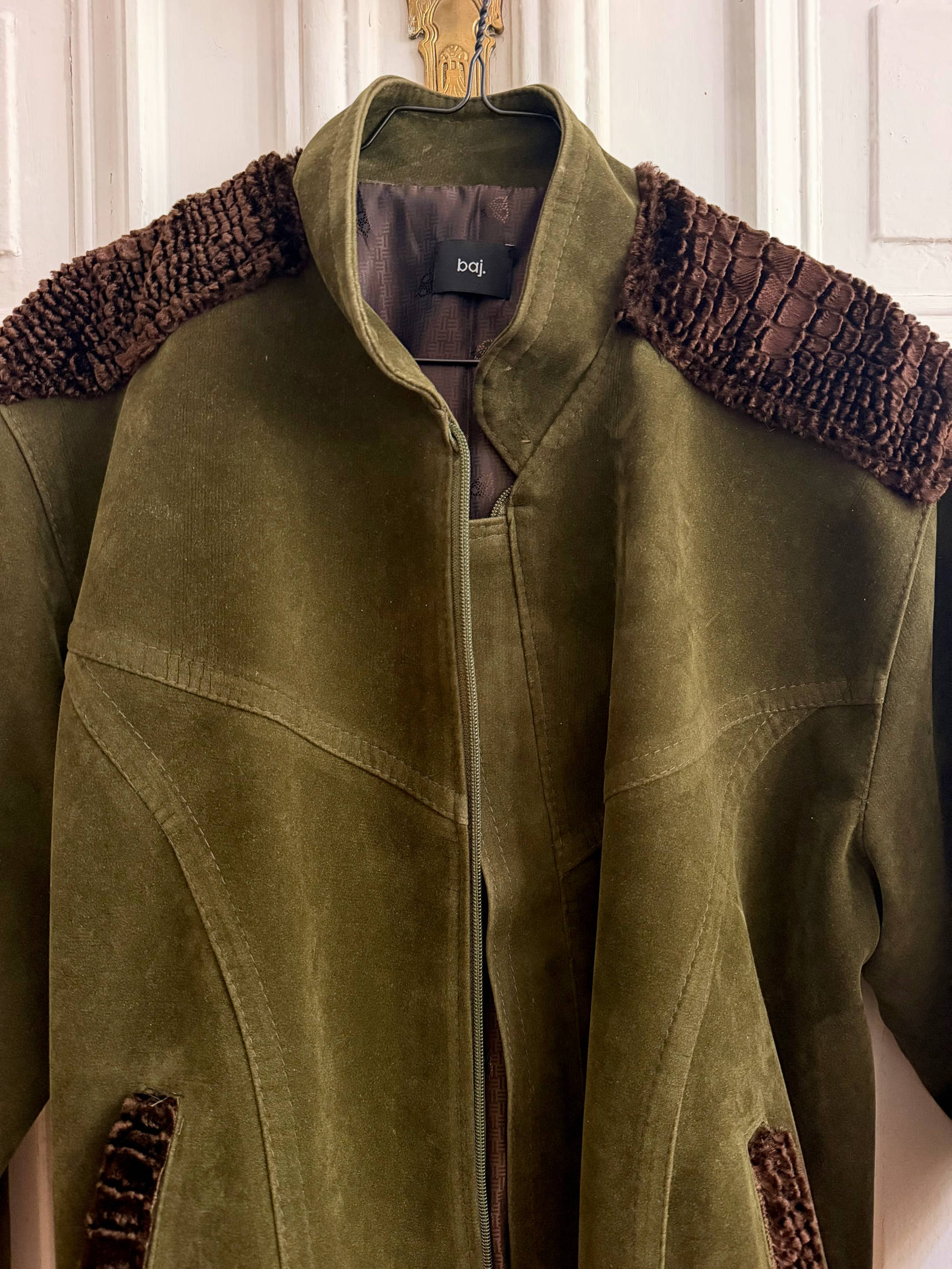 An olive green suede jacket with brown textured shoulder yokes and matching brown textured pocket trims, hanging on a wooden hanger against a white paneled door.