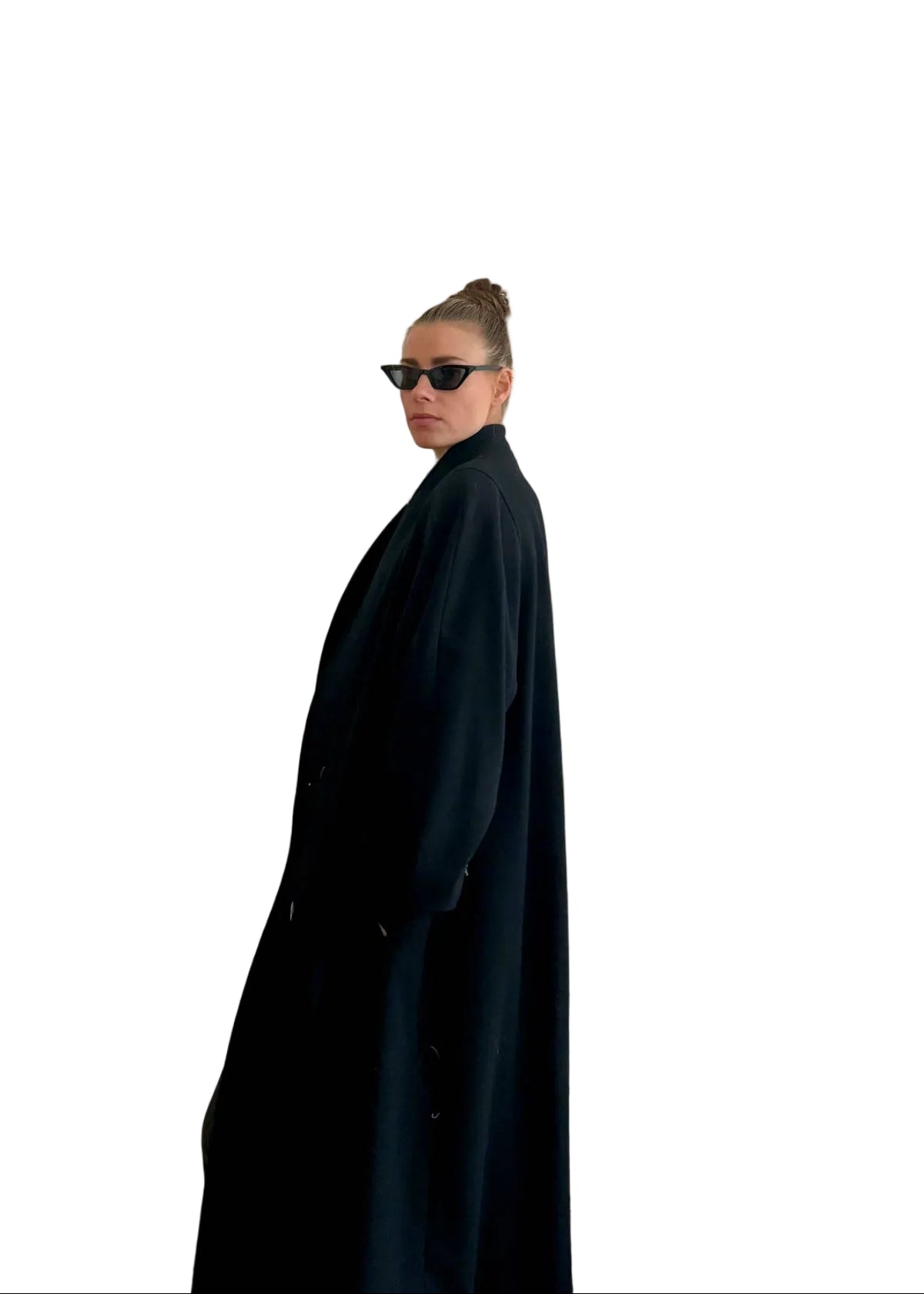 A person wearing a long black coat and black sunglasses stands in profile in front of a white paneled door in an indoor room.