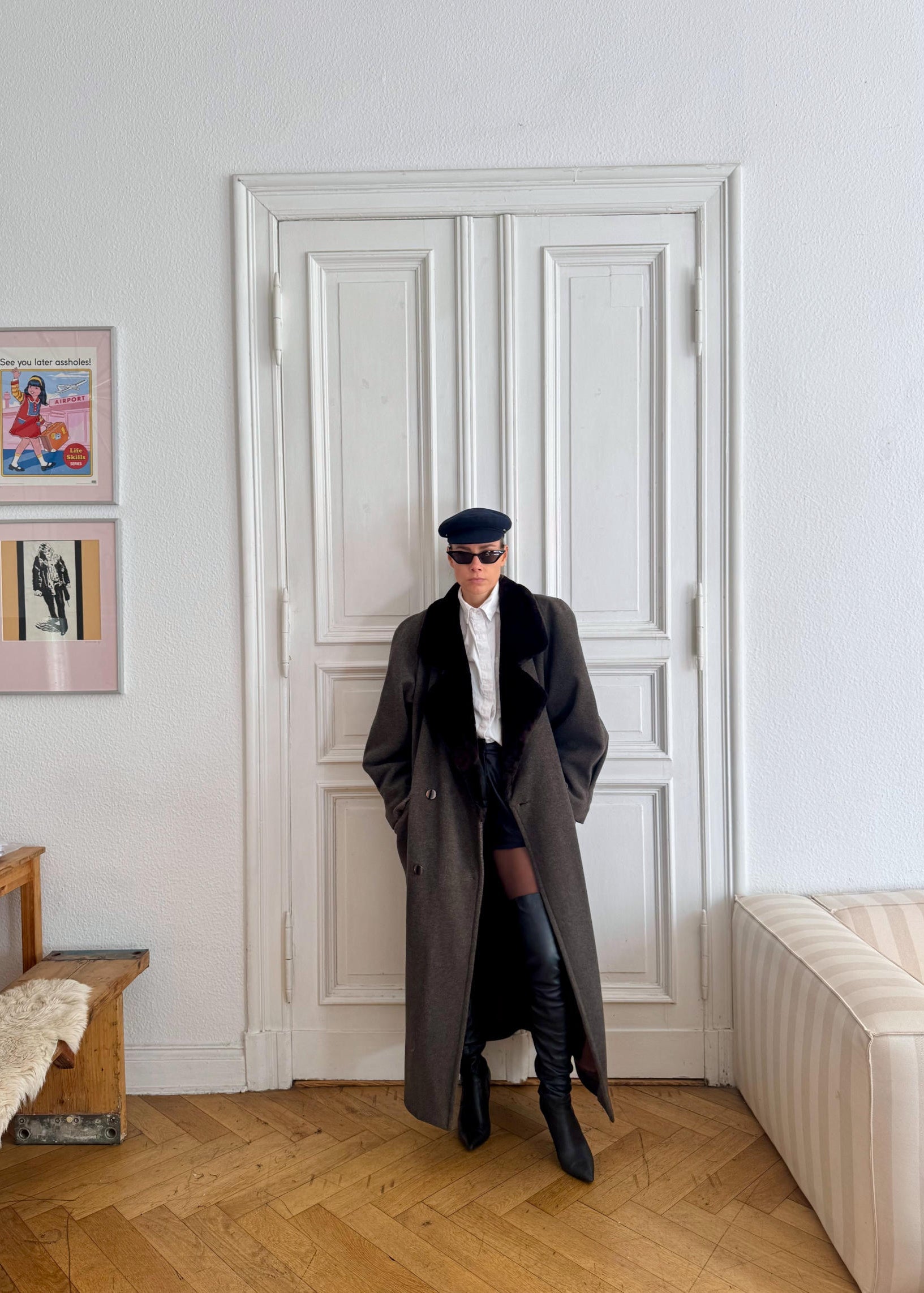 Person wearing a long coat and black boots standing in a room with a white door and wooden floor.