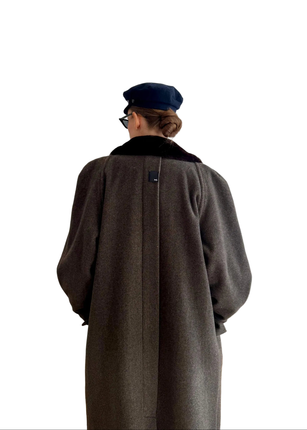 Person wearing a long coat and cap standing in a room with white walls and wooden floor.