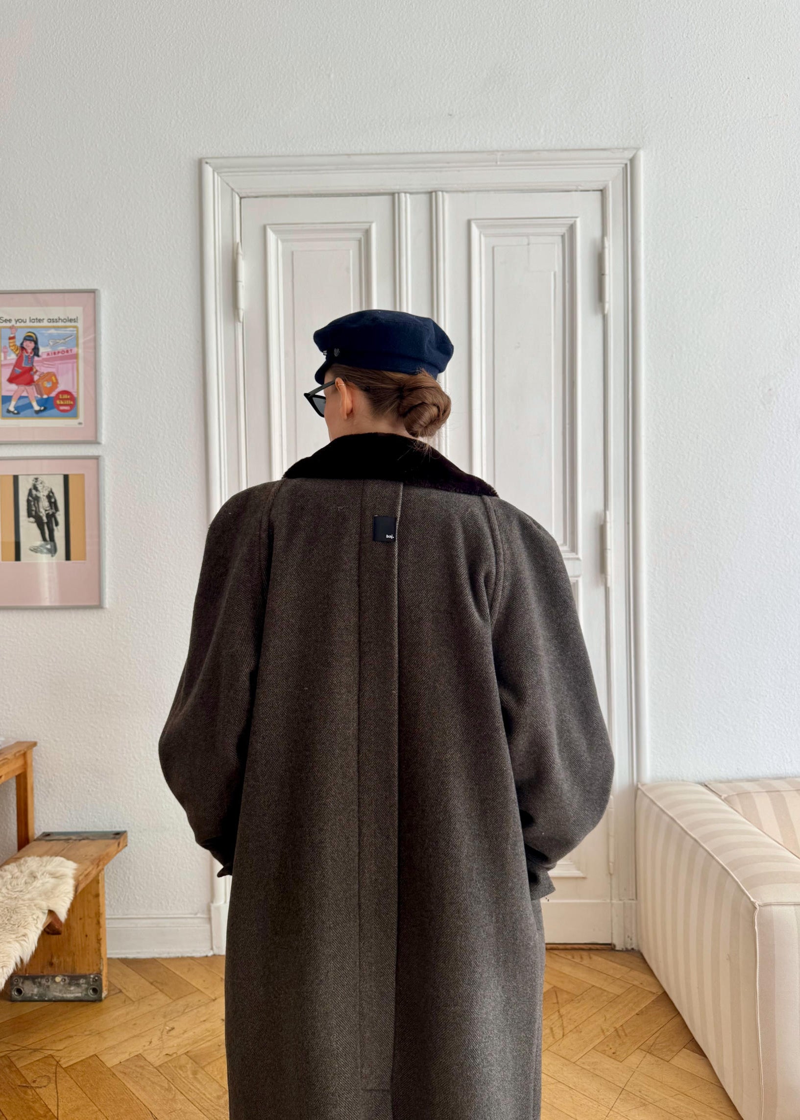 Person wearing a long coat and cap standing in a room with white walls and wooden floor.