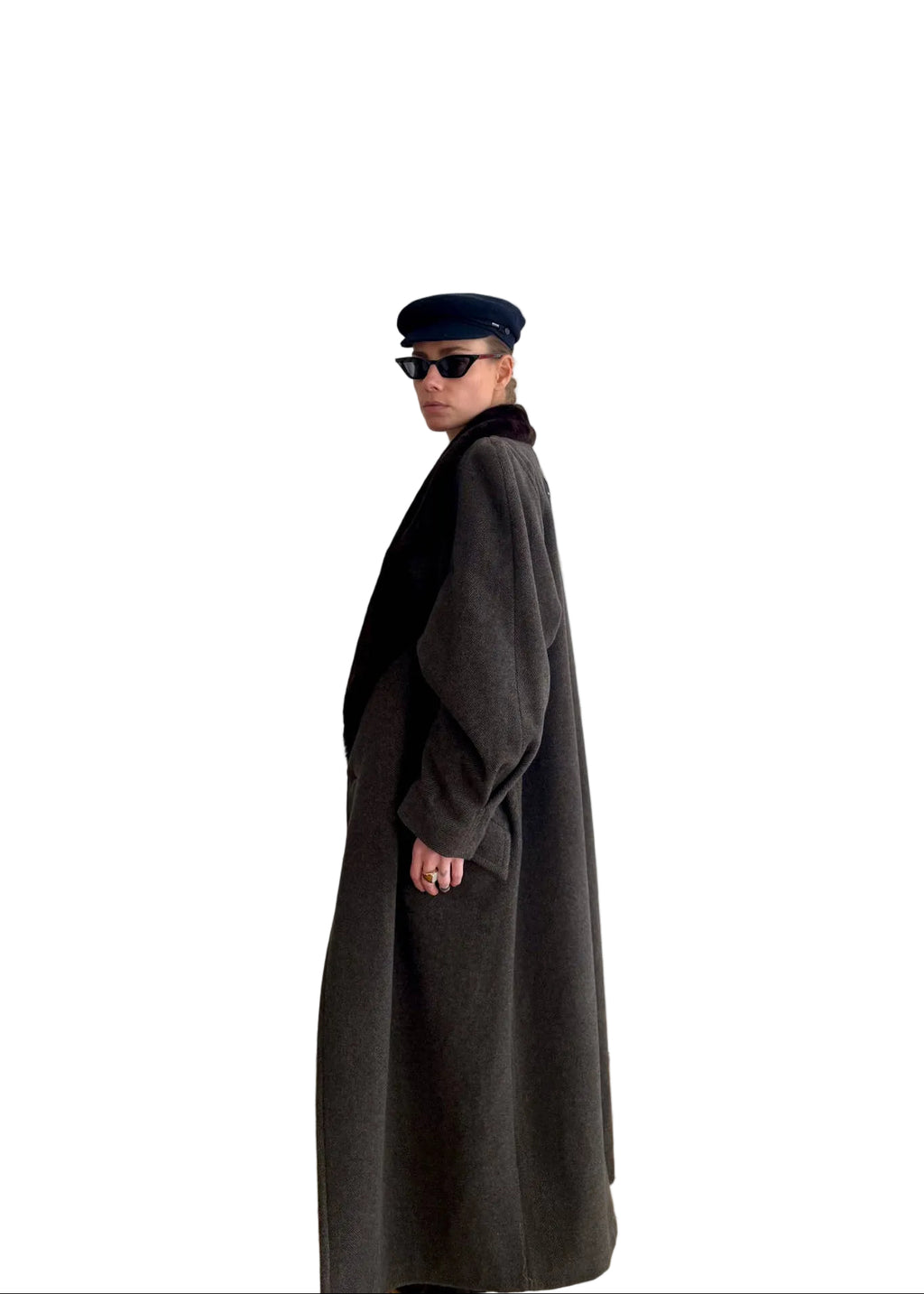 Person wearing a long dark coat and sunglasses standing in a room with a white door and framed pictures on the wall.