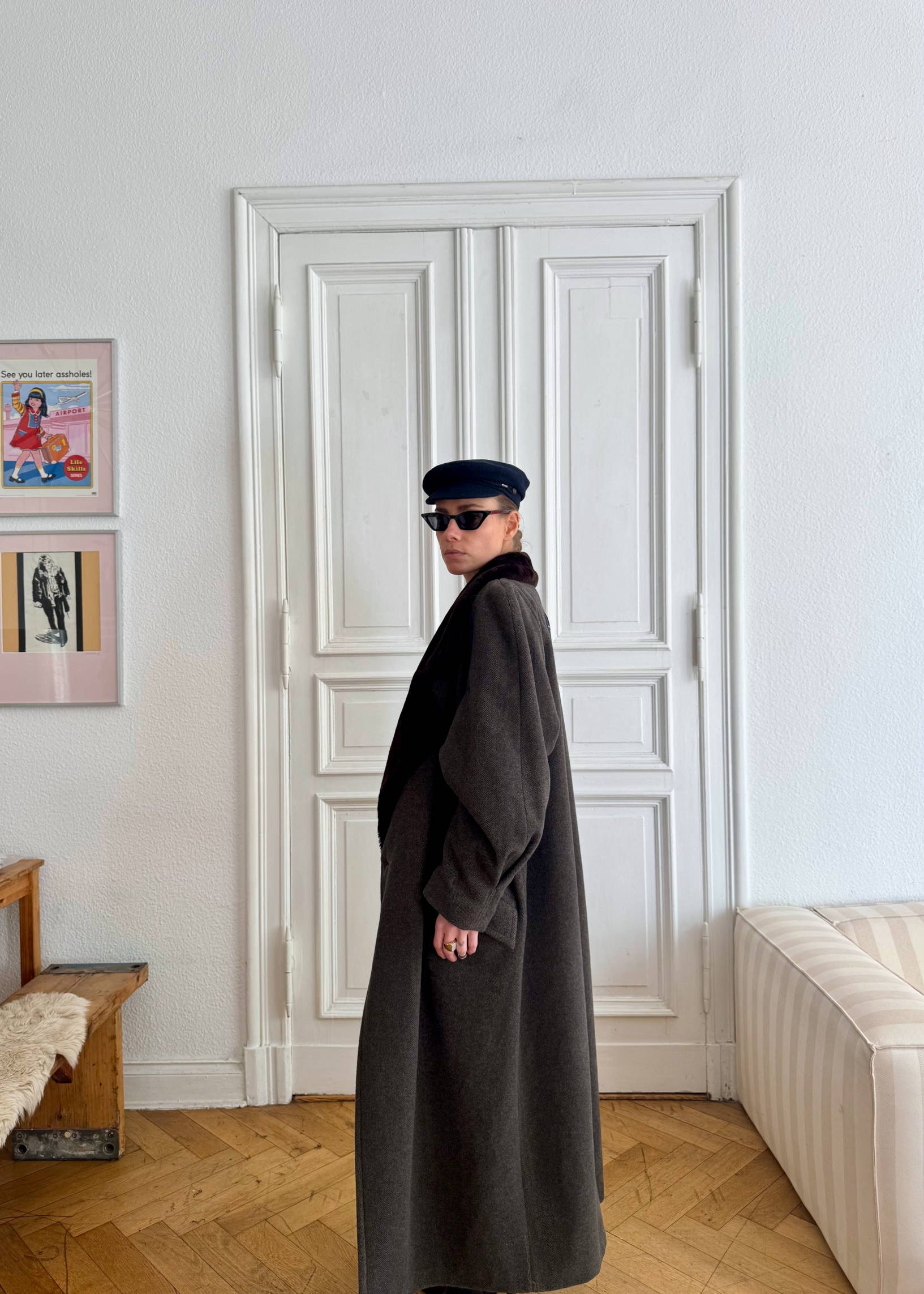 Person wearing a long dark coat and sunglasses standing in a room with a white door and framed pictures on the wall.