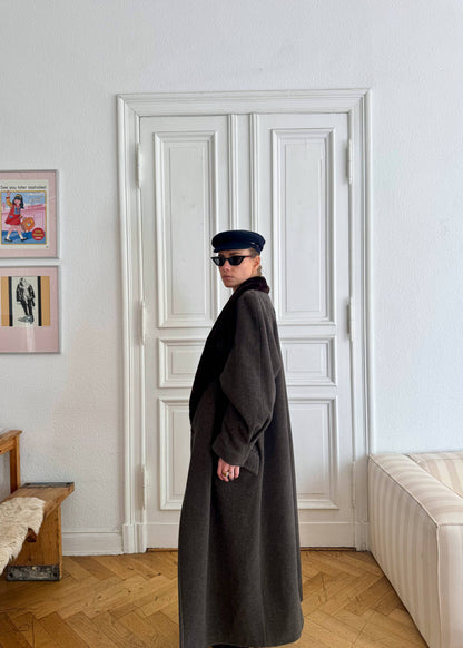 Person wearing a long dark coat and sunglasses standing in a room with a white door and framed pictures on the wall.