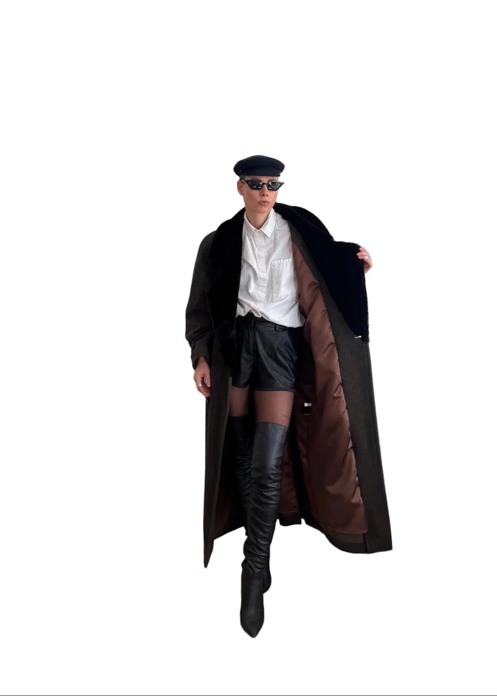 Person standing in a room with a white door, wearing a long coat, white shirt, black skirt, and black knee-high boots.