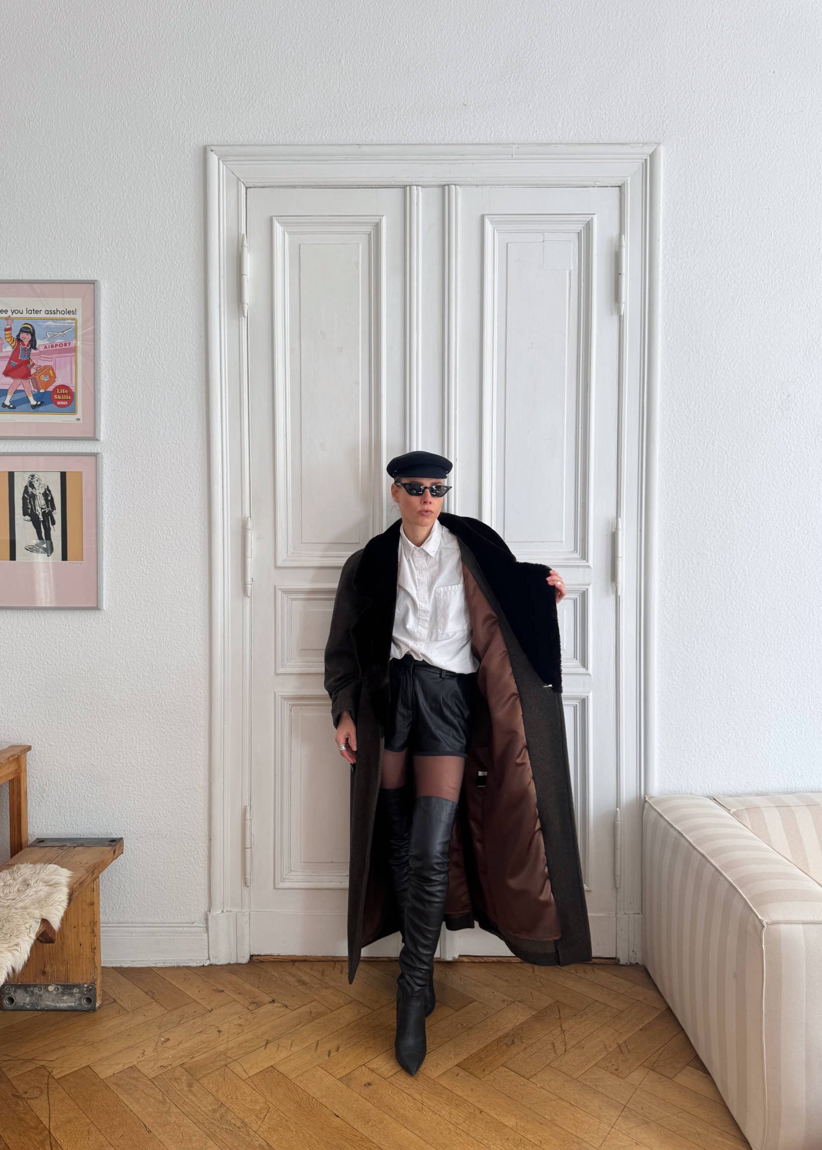 Person standing in a room with a white door, wearing a long coat, white shirt, black skirt, and black knee-high boots.