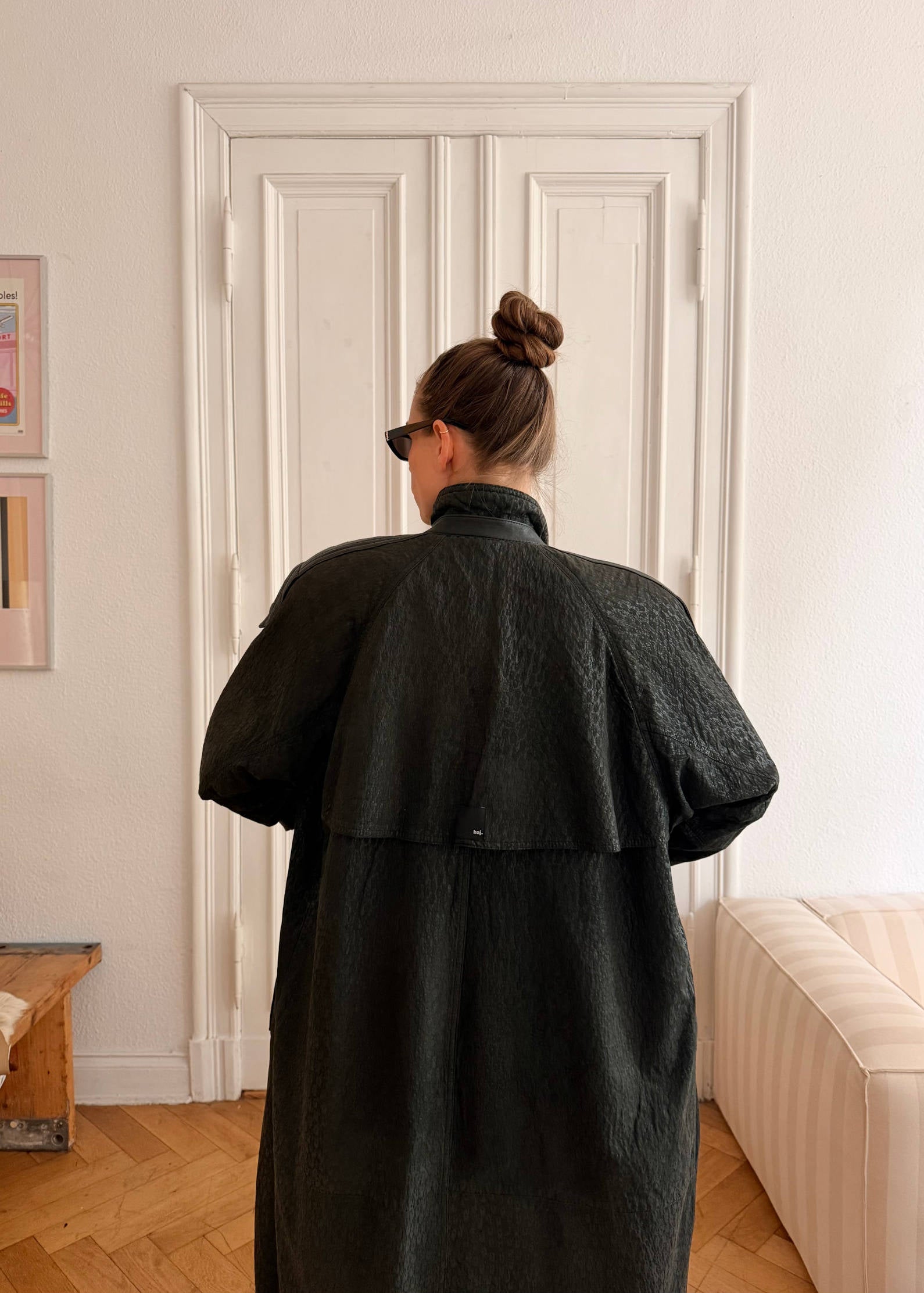 Person wearing a black coat standing in a room with white walls and wooden flooring.