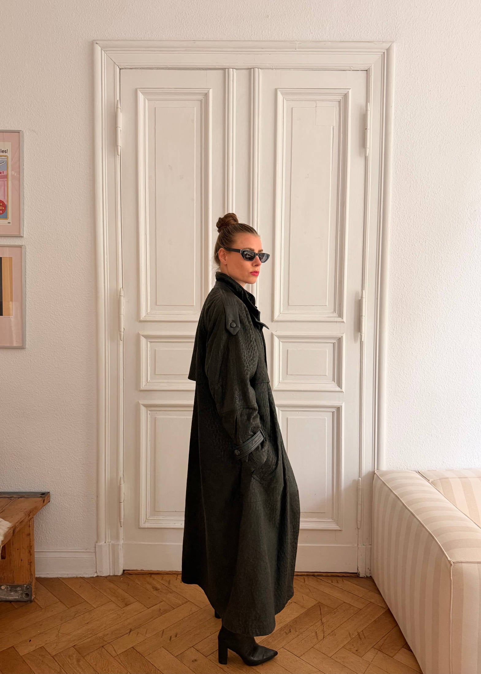Person wearing a long dark coat standing in a room with a white door and wooden floor.
