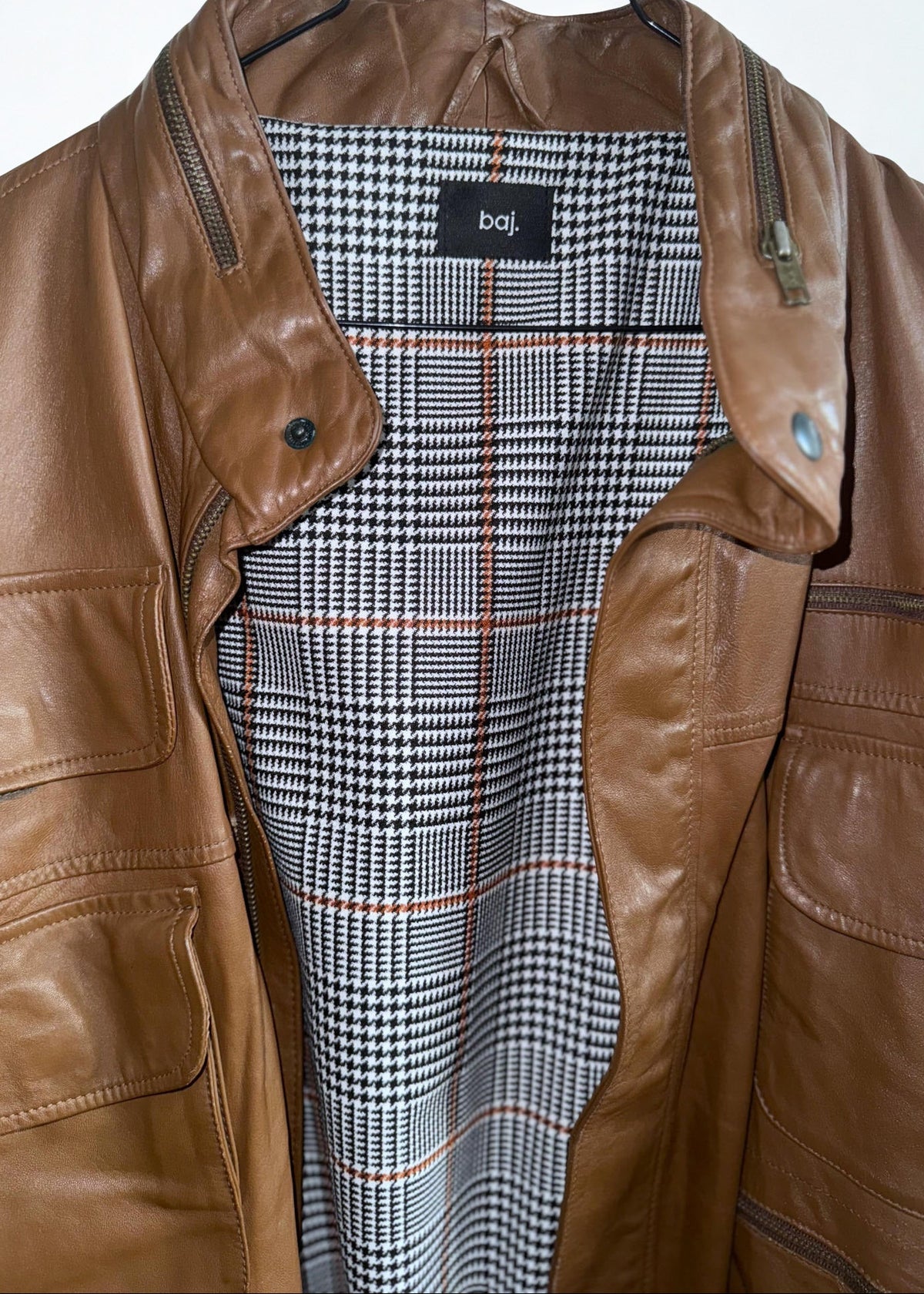 A brown leather jacket with a black and white houndstooth patterned lining, hanging on a black hanger against a white background.
